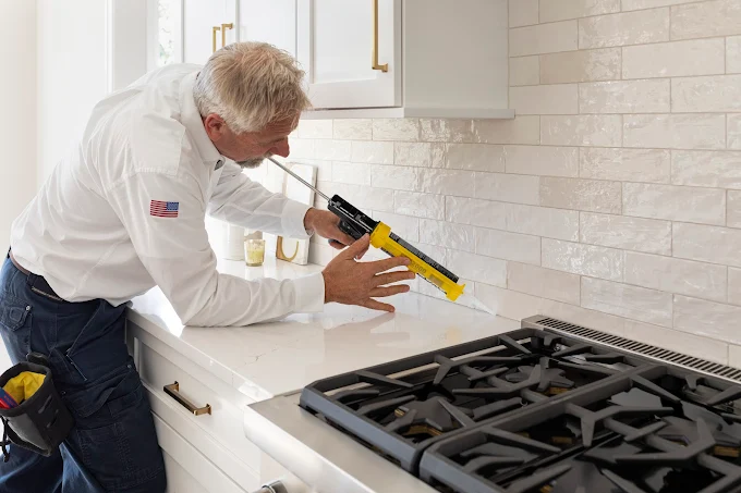 A handyman applies caulk along a kitchen countertop backsplash, sealing the edge between the tile and counter. He is wearing a white uniform with an American flag patch and tool belt.