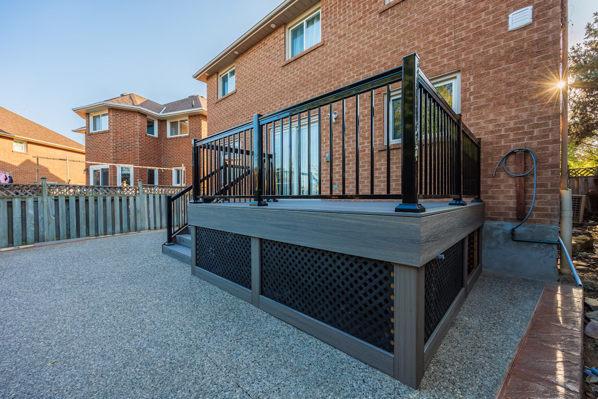 Modern raised wooden deck with black metal railing attached to a red brick house, featuring lattice skirting and textured concrete patio.